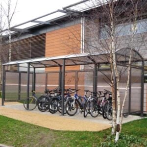 Abri pour vélos intérieur sur dalle de béton, structure métallique et toit transparent, situé près d’un bâtiment avec végétation autour.