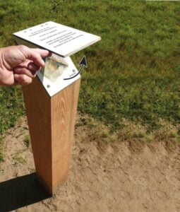 Borne Polyèdre en bois avec plaque d’information vissée, actionnée par une main devant un espace extérieur.