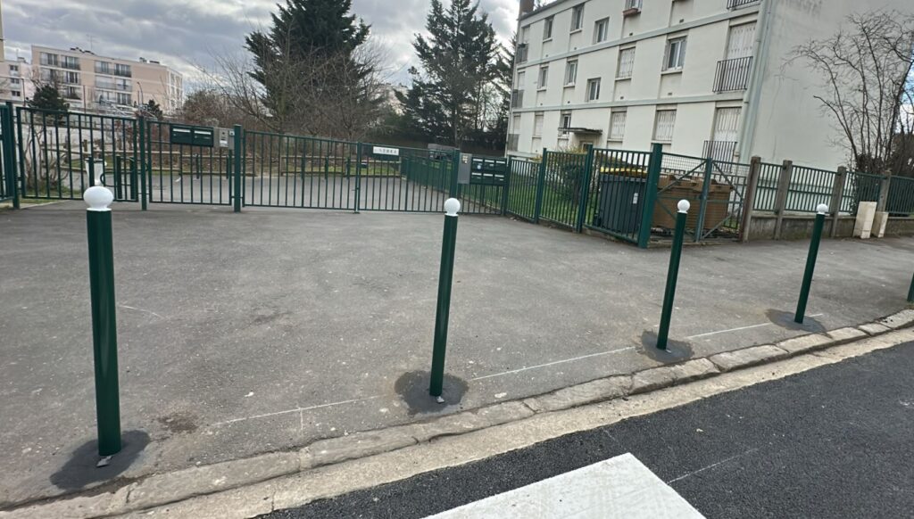 Potelets urbains boule vert foncé avec capuchon blanc sur trottoir, délimitant et sécurisant un espace pavé devant un parking et une clôture métallique.